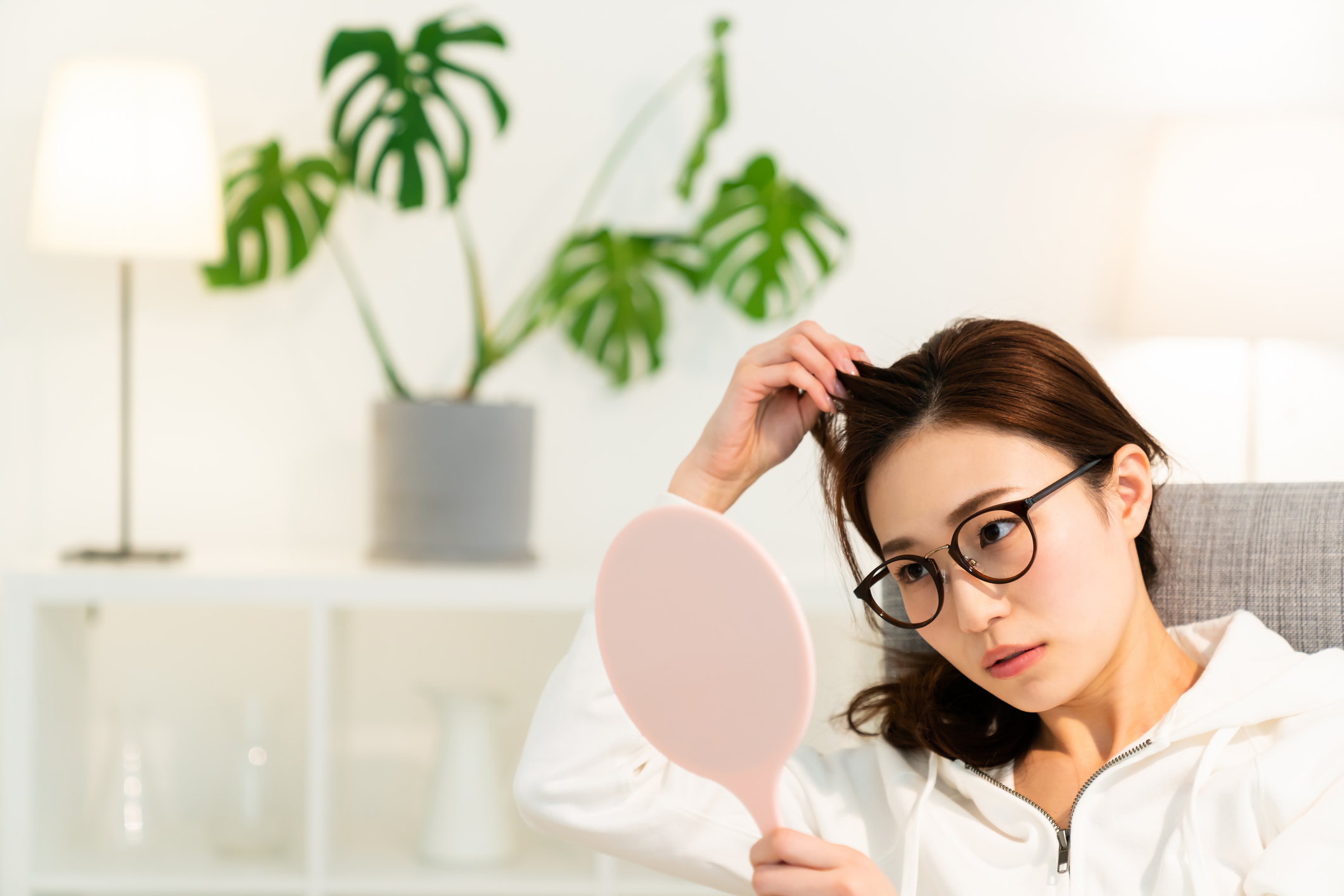 Concerned Asian woman with long brown hair examines dry scalp in handheld mirror.