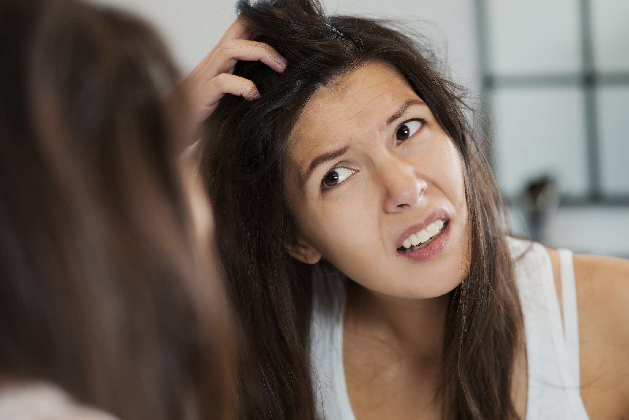 Woman with long brown hair scratches scalp while looking in the mirror.